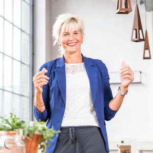 lachende vrouw bij groot raam, blond haar en blauw jasje. Ze lacht en is relaxt.