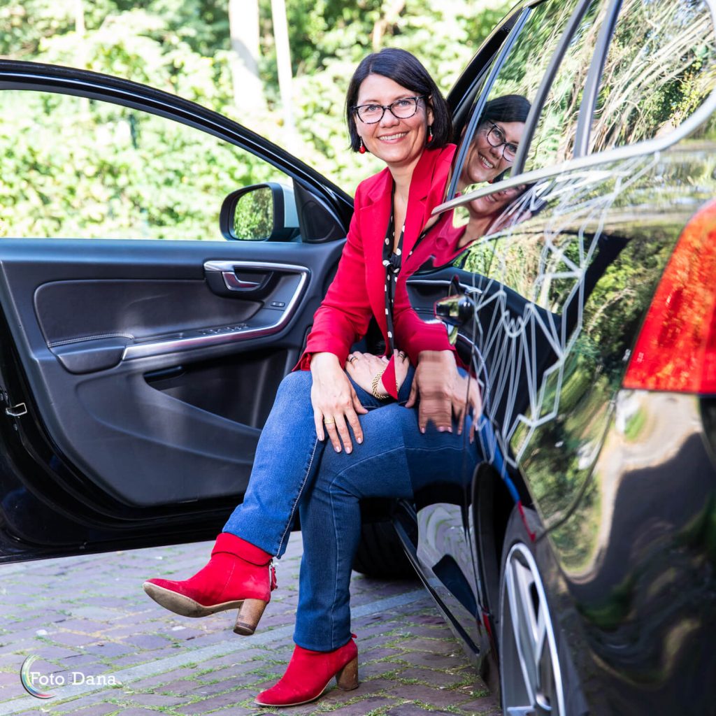 Buitenportret vrouw met halflang zwarthaar en bril op zit half in auto. rode laarsjes en rode jas